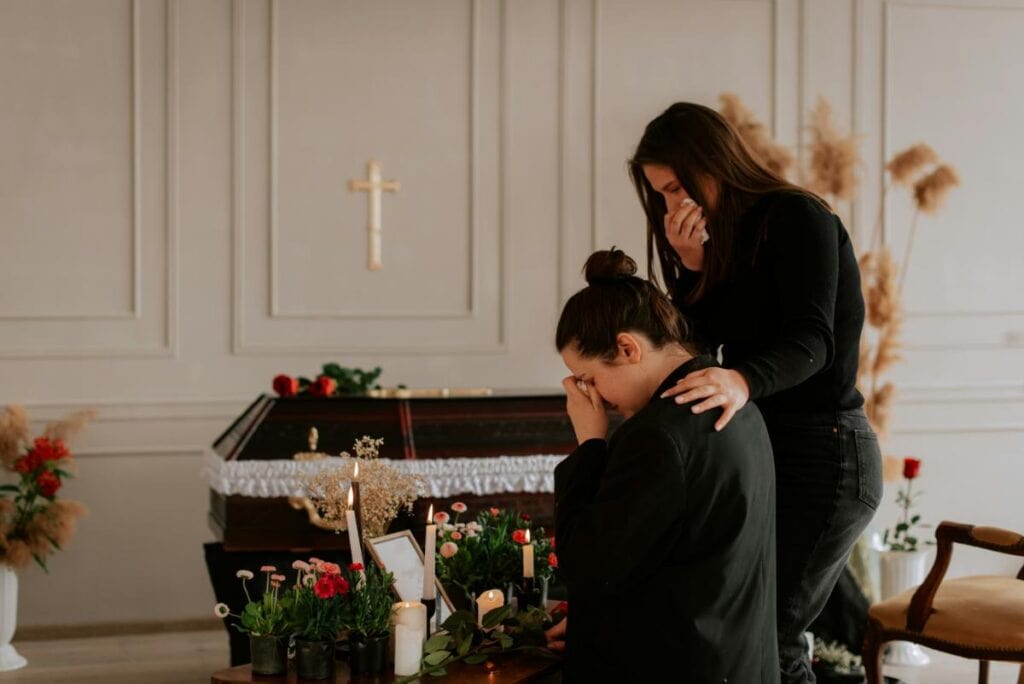 Two women mourn beside a flower-lined casket during a funeral; one kneels in tears while the other offers comfort with a gentle hand on her shoulder.