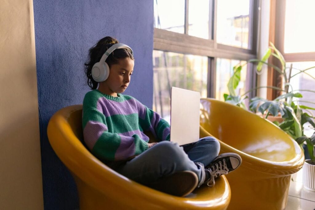 Young girl wearing headphones and a striped sweater, sitting cross-legged in a modern yellow chair while focused on a laptop screen.