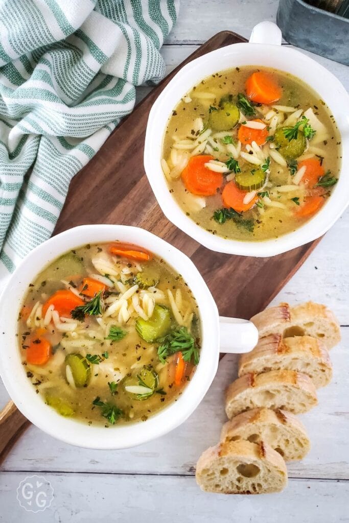 Two bowls of homemade chicken orzo soup filled with carrots, celery, and pasta, served on a wooden board with sliced bread on the side.