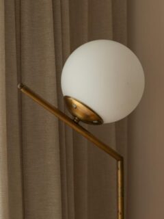Detail of a brass floor lamp with a frosted globe shade positioned in front of textured beige curtains in a softly lit interior.