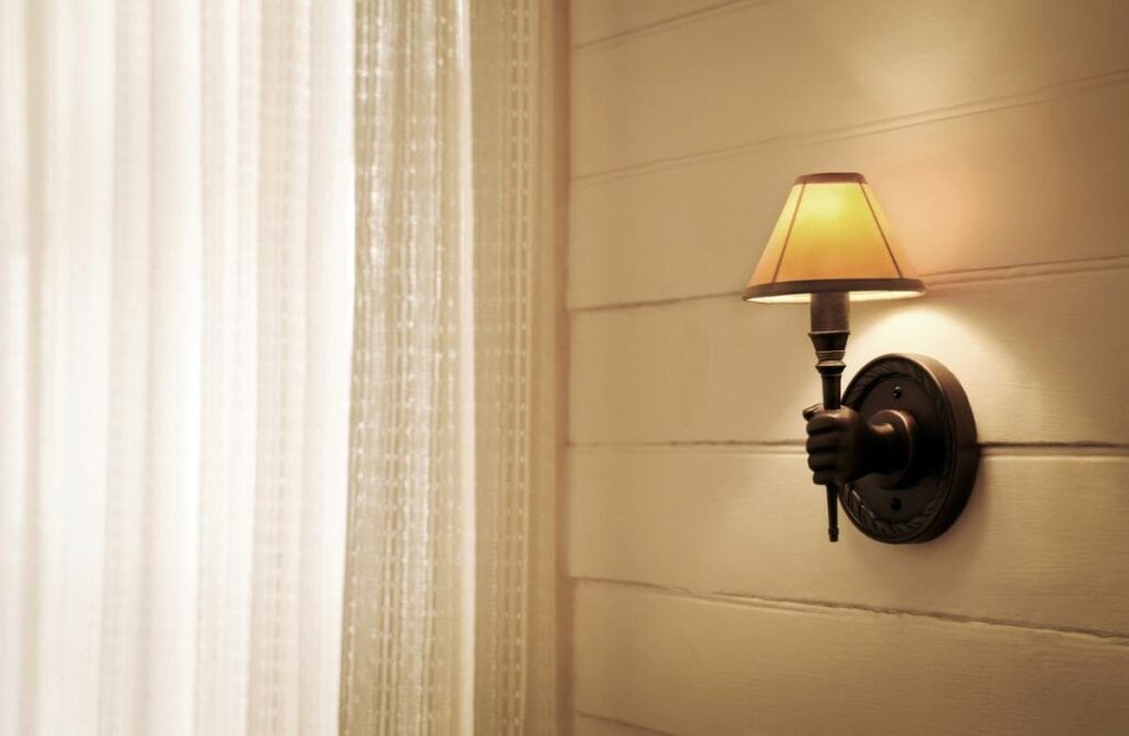 Wall sconce shaped like a hand holding a small lamp with a warm-toned fabric shade, mounted on a cream-colored wooden wall beside sheer curtains.