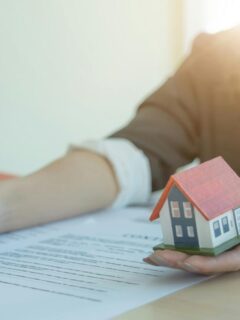 a woman holding a model of a house and using a pen to point to a contract signature line