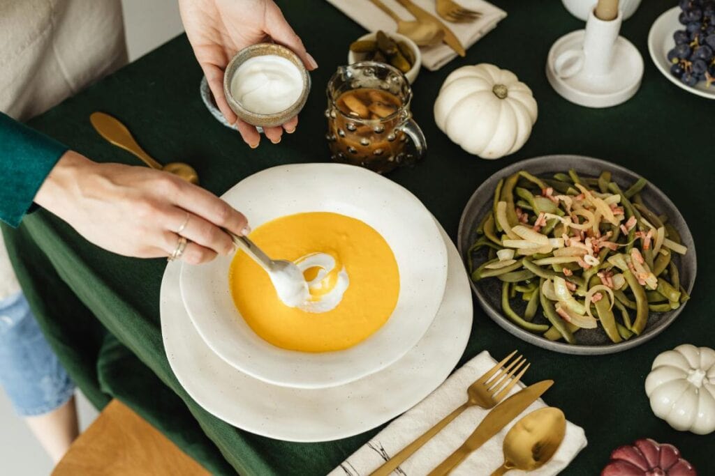 Creamy pumpkin soup being topped with yogurt at a cozy dinner table with seasonal vegetables and autumn decor.