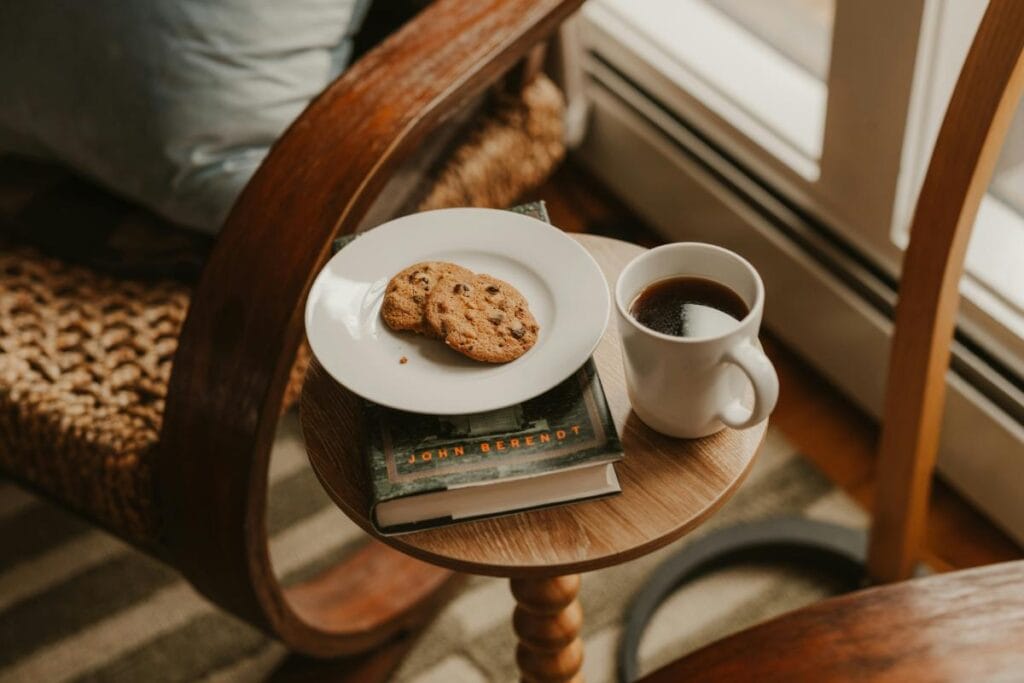 Cozy coffee and cookie on a small wooden side table beside a reading chair, creating a relaxed caf&eacute;-style home atmosphere.