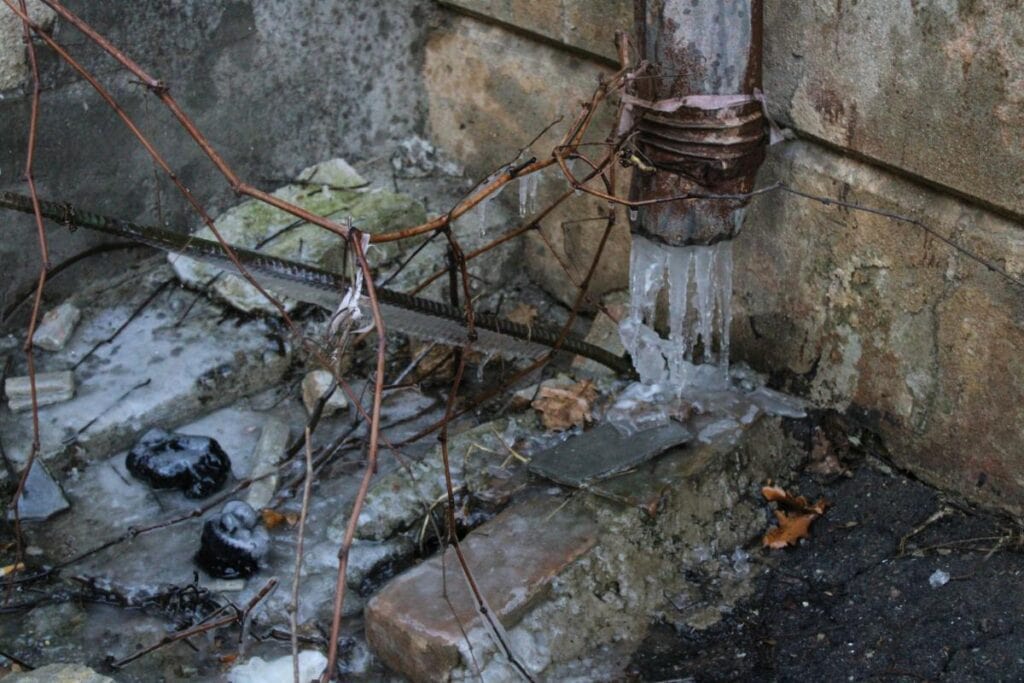 Outdoor pipe frozen with ice buildup along a house foundation during winter.