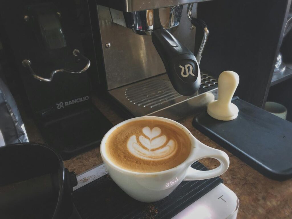 Fresh latte with latte art under a home espresso machine and coffee grinder, representing a caf&eacute;-style drink station setup.