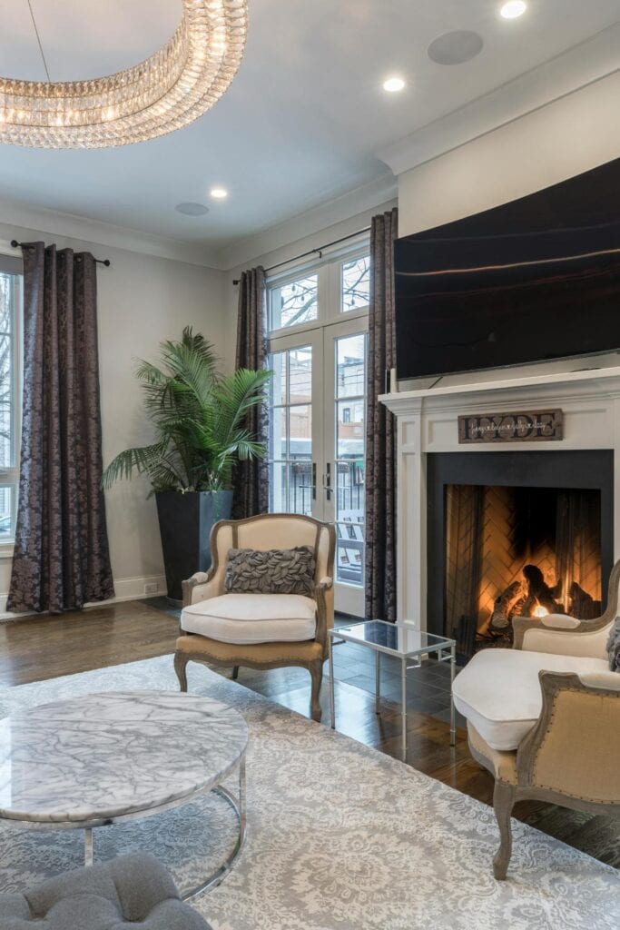 Elegant living room with a fireplace, marble coffee table, armchairs, crystal chandelier, and large windows creating a bright, sophisticated space.