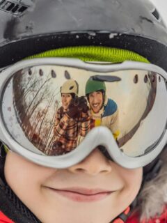 smiling kid in ski goggles with parents showing in reflection of goggles