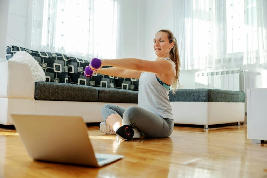 oman exercising at home with dumbbells while following a workout video on a laptop