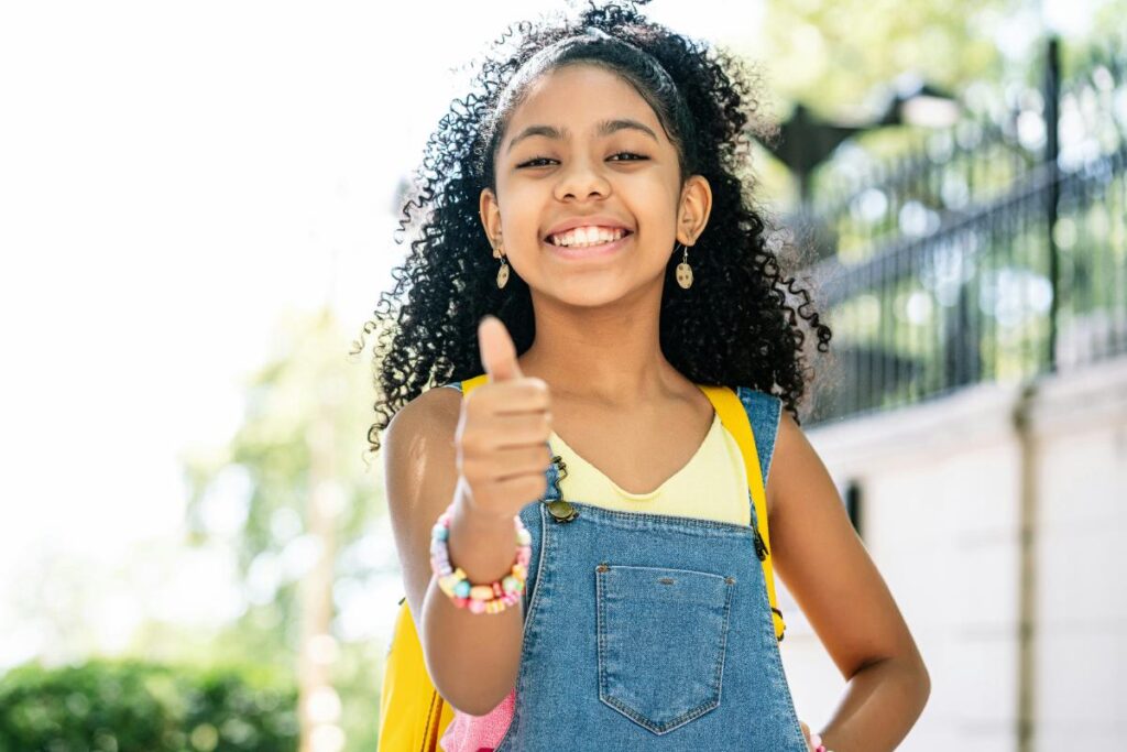 Smiling young girl with backpack giving thumbs up outdoors, representing confidence, independence, and positive self-esteem