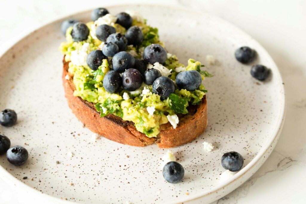 Avocado toast topped with fresh blueberries and crumbled cheese on a white plate, highlighting a healthy and balanced breakfast option.