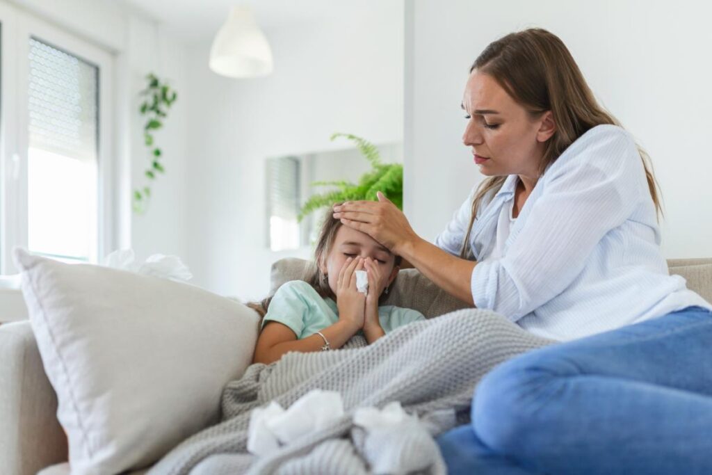 mom curled up on the couch with her sick daughter, checking her forehead temperature with her hand