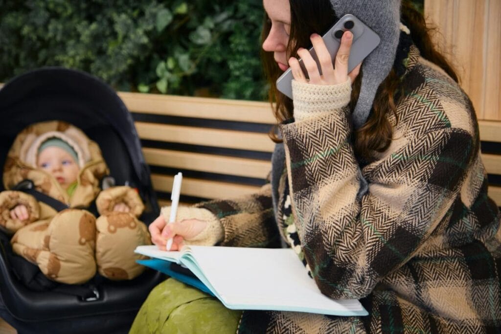 Mom talking on her phone while writing notes beside a stroller, showing how busy parents stay organized while out with a baby.