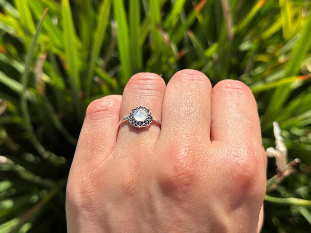 Delicate silver opal ring worn on a hand outdoors, showcasing the gemstone's subtle iridescence against a natural green background.