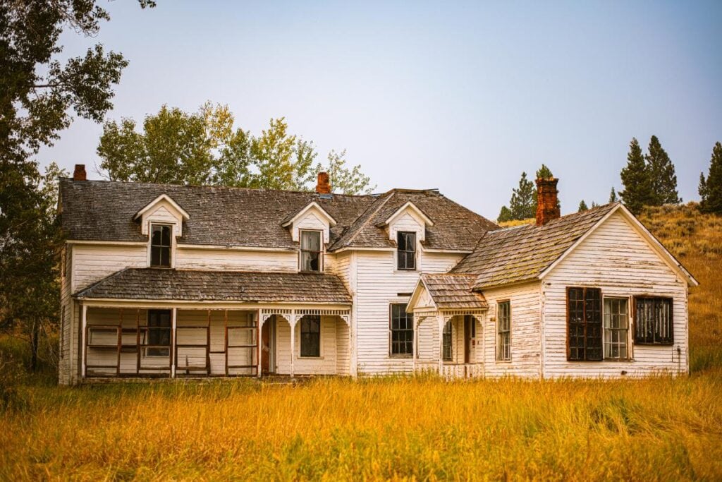 Abandoned white farmhouse with weathered siding and overgrown grass, representing an unwanted property ready for cleanup or sale.