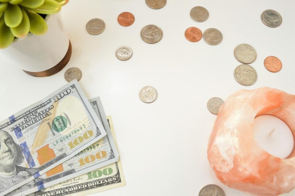 U.S. cash and coins arranged on a white desk beside a plant and candle, representing budgeting, saving money, and personal finance planning for creative entrepreneurs.
