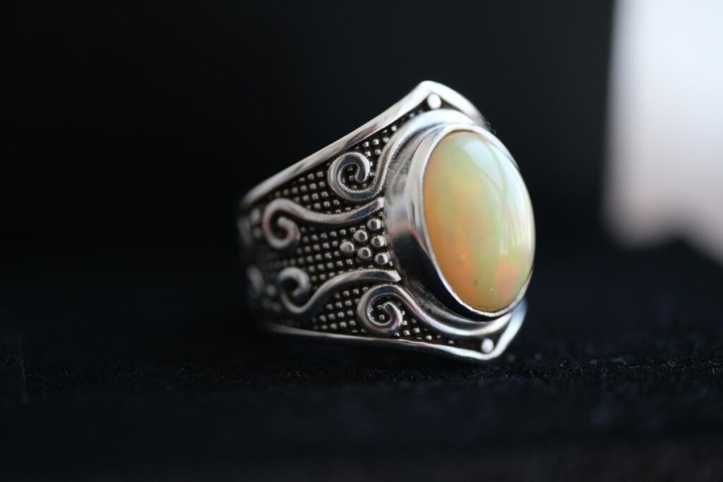 Vintage-style silver opal ring with intricate detailing and a luminous oval gemstone, photographed against a dark background.