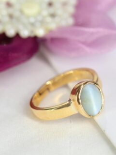 Minimalist gold opal ring with an oval cabochon stone displayed beside soft pink and purple flowers on a white surface.
