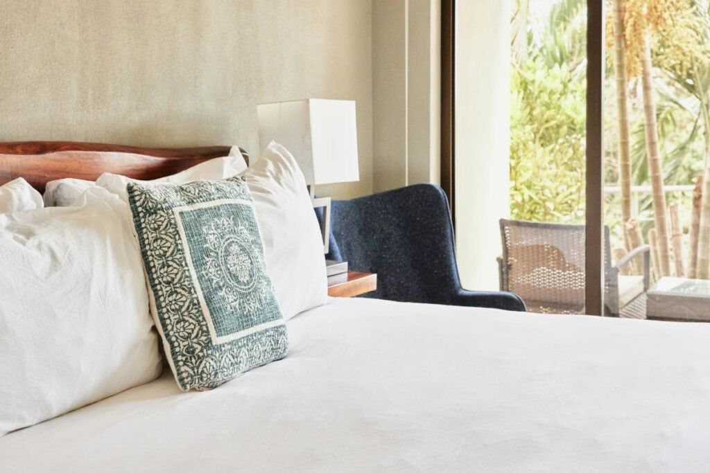 Bright and inviting bedroom with a neatly made bed, white bedding, decorative accent pillow, and natural sunlight streaming through large windows.
