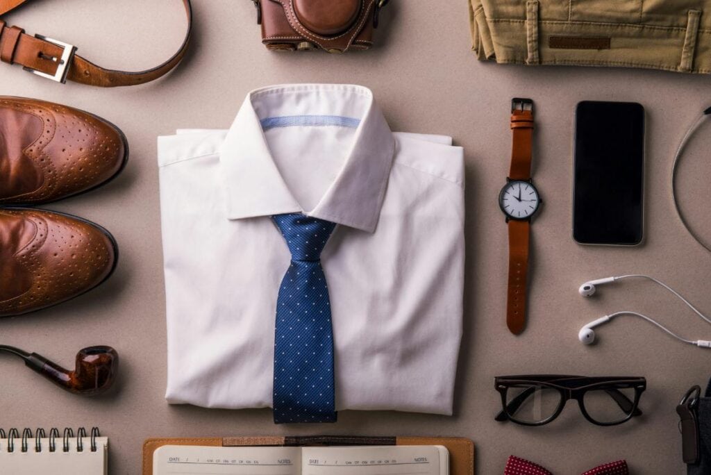  Flat lay of men's wardrobe essentials with a dress shirt, tie, watch, belt, shoes, and everyday accessories.