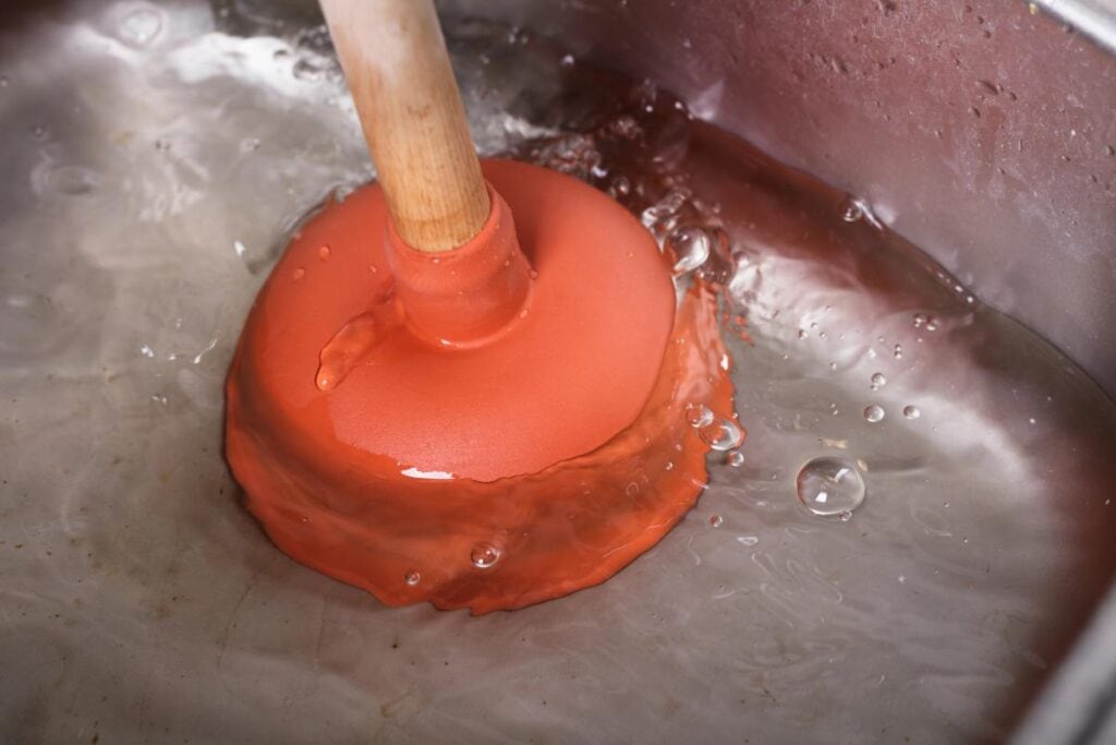 Plunger clearing a clogged kitchen sink with standing water, illustrating an early warning sign that a home may need professional drain cleaning.