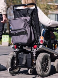 Power wheelchair user traveling outdoors with a companion on a sunny day.