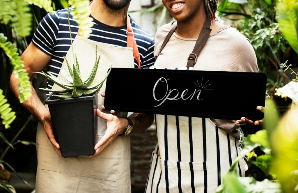 Small business owners holding an open sign and potted plant in a garden shop