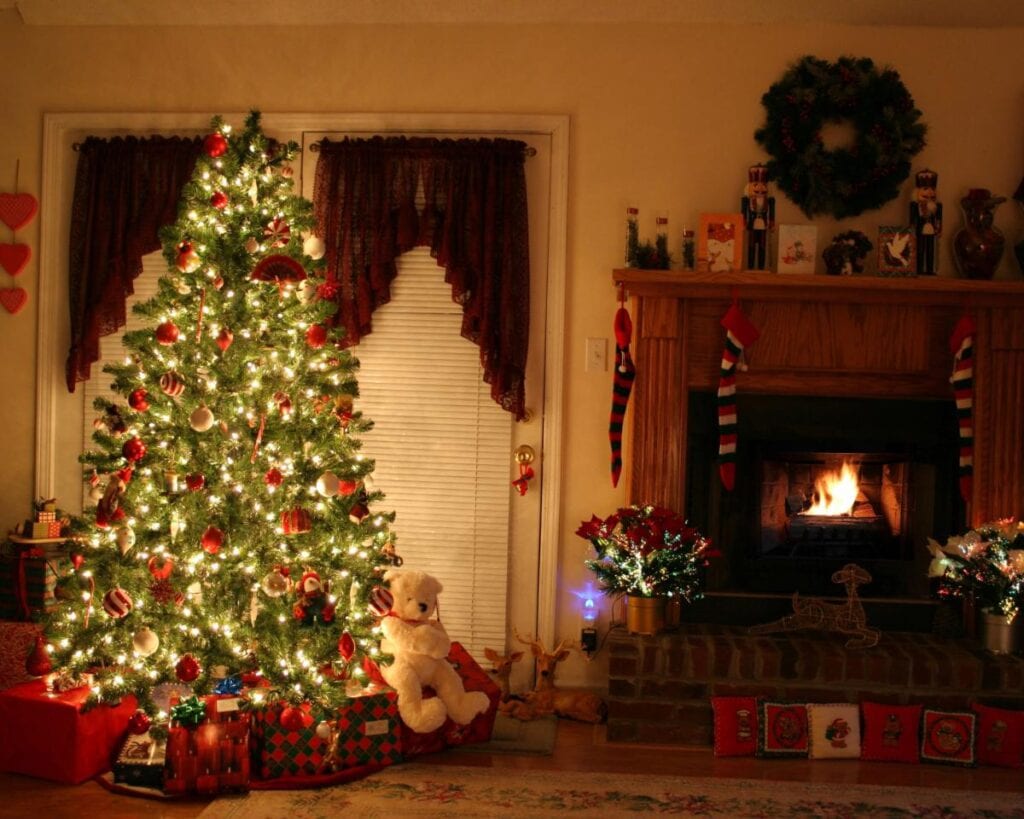 Nostalgic Christmas tree glowing with warm lights and red ornaments beside a fireplace, wreath, stockings, and wrapped gifts.