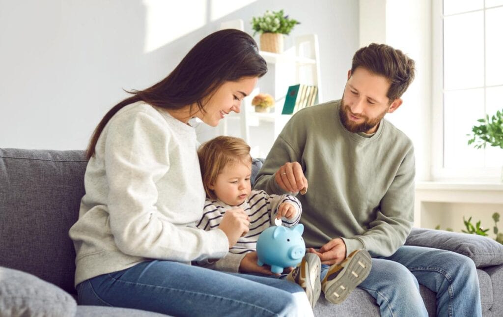 Parents helping their young child put money into a piggy bank at home