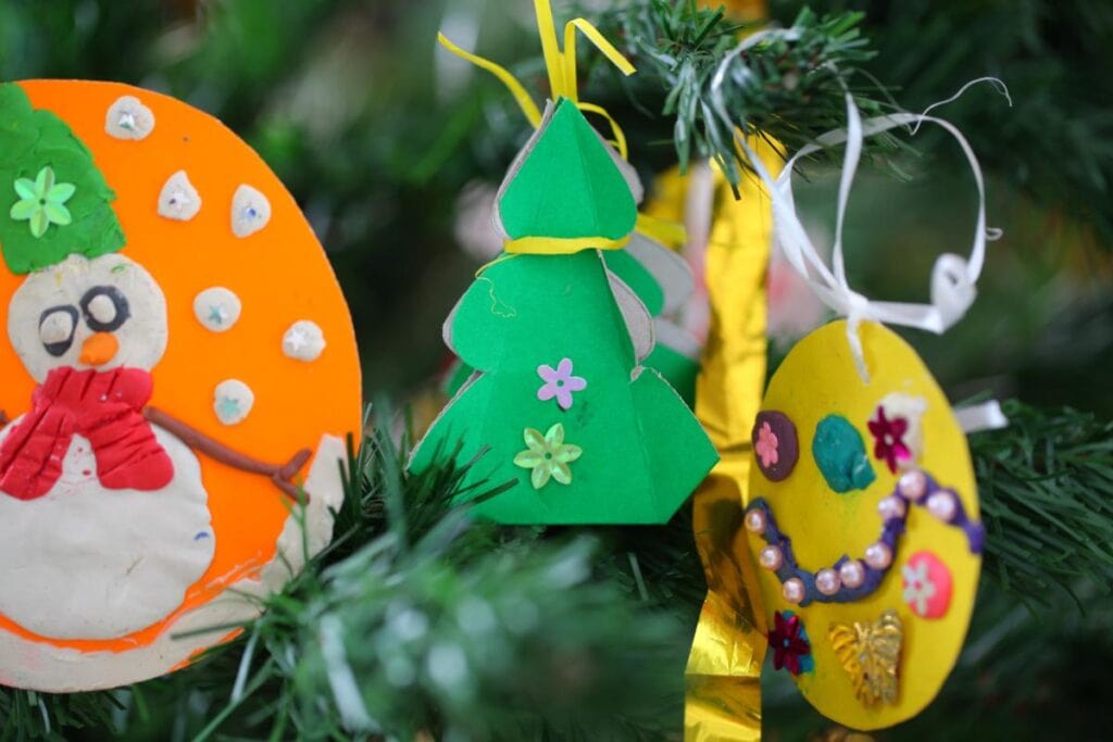 Colorful handmade Christmas ornaments, including a snowman, paper tree, and decorated craft ornament, hanging on evergreen branches.