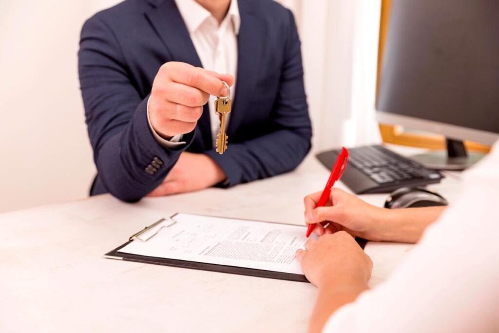 Real estate professional handing over keys while a client signs property documents at a desk during a commercial closing.