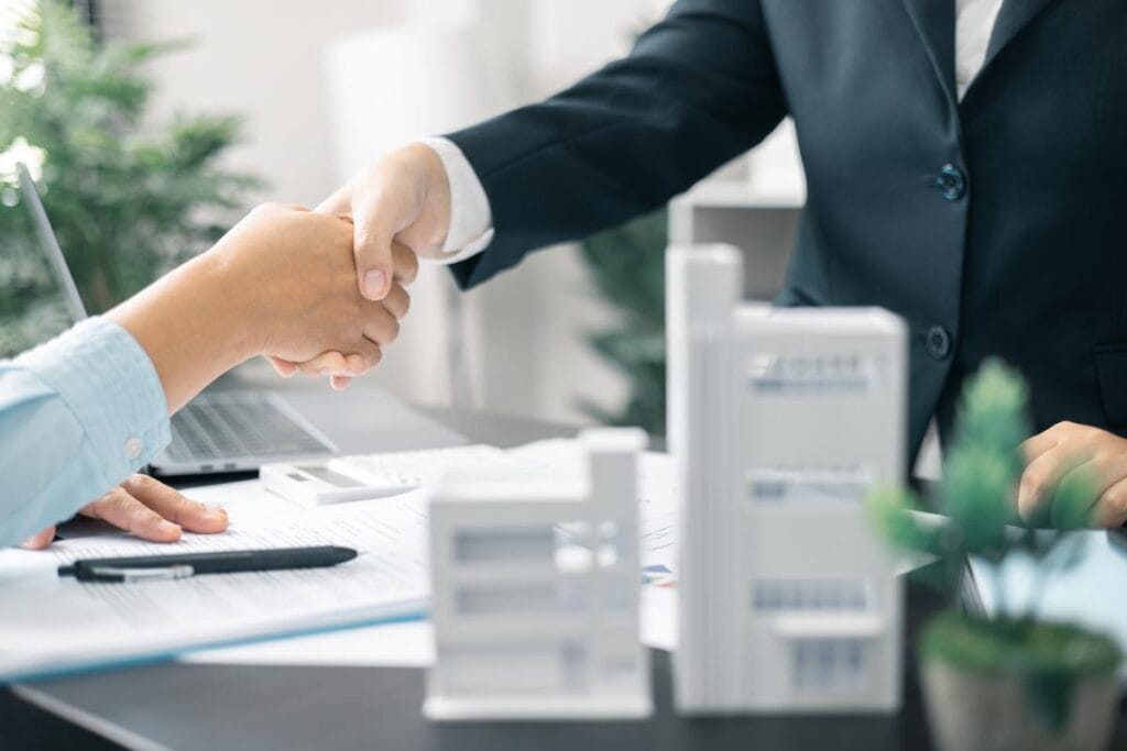 Two business professionals shaking hands across a desk with building models and closing documents in the foreground.