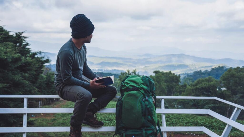 young man sitting on a fence to take a break from backpacking to read